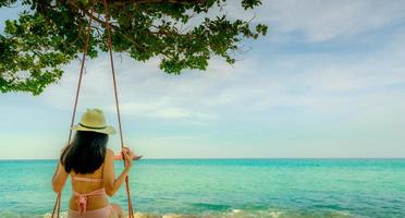 mujer asiática usa traje de baño y sombrero columpia los columpios en la playa de arena y luce un hermoso paraíso tropical mar y cielo en un día soleado. vacaciones de verano. vibras de verano. disfrutando y relajando a una chica de vacaciones foto