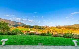 campo de hierba verde en la colina. paisaje de pueblo en el valle. bosque de pinos en la montaña con cielo azul y nubes blancas en un día soleado en europa. asiento vacío en el jardín del parque. pequeña ciudad en el valle foto