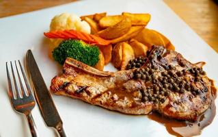 Steak topped with sauce and pepper serve with potato fried and carrots, cauliflower and broccoli steamed. Steak on white plate with stainless steel fork and knife put on wooden table. Tomahawk steak. photo