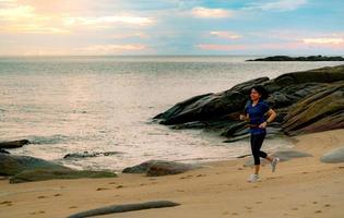 mujer asiática corriendo en la playa tropical por la mañana con un hermoso cielo de amanecer. entrenamiento al aire libre de mujer sana en forma. La chica en forma usa banda inteligente y riñonera. mujer corriendo para hacer ejercicio cardiovascular. foto