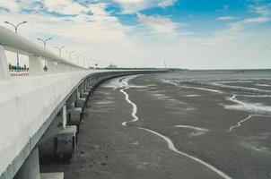 Curved coastal concrete highway road with lower water in the mud beach with blue sky and clouds. Road trip travel. Summer journey at tropical Cement bunting bridge. Nature abstract background. photo