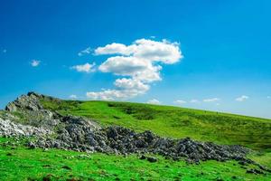 paisaje de hierba verde y colina rocosa en primavera con hermoso cielo azul y nubes blancas. campo o vista rural. fondo de naturaleza en un día soleado. ambiente de aire fresco. piedra en la montaña. foto