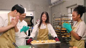 Young Asian female cooking class student brings tray of baked pies from electric oven, senior chef beside, happy pastry cuisine in culinary course lesson, food occupation in stainless steel kitchen.