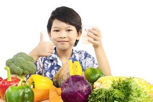 niño sano asiático que muestra una expresión feliz con un vaso de leche y una variedad de verduras frescas y coloridas sobre fondo blanco foto