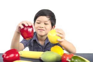 Chico sano asiático mostrando expresión feliz con variedad de frutas y verduras de colores sobre fondo blanco. foto