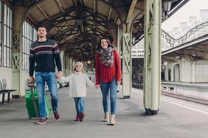 una foto de una familia feliz que va a tener un buen viaje durante las vacaciones, llevar una bolsa, caminar en la plataforma de la estación de tren, estar de buen humor. padre, madre e hijo llegan de viaje. concepto de viaje