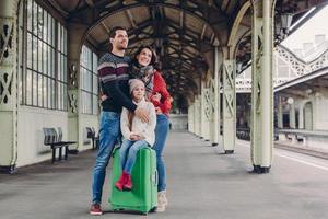 Pleased wife and husband cuddle with love, their daughter sits at suitcase, pose together on platform, look in ditance, wait for train, have pleased facial expressions. Happy family travel abroad photo