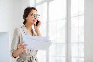 Pensive businesswoman stands sideways to camera, holds paper documents, has phone talk, prepares financial report, poses in coworking space, wears optical glasses and formal suit. Paperwork. photo