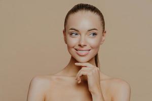 Smiling well groomed young woman with beauty face care, healthy glowing skin, touches chin gently and looks aside, poses shirtless against brown studio background, loves her skin, feels refreshed photo