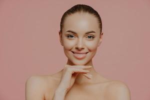 Pretty young European woman touches chin gently, enjoys flawless of skin after beauty procedures, poses nude, has natural makeup, isolated on pink background. Beauty, spa and wellness concept photo