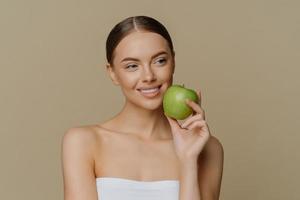 una mujer europea encantadora y pensativa sostiene manzana cerca de la cara sonríe suavemente tiene dientes blancos perfectos piel sana y limpia envuelta en una toalla de ducha se para con los hombros desnudos contra un fondo marrón. foto