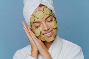 Relaxed satisfied woman tilts head on palms pressed together keeps eyes shut and smiles gently makes facial mask applies cucumber slices on face enjoys morning skin care routine poses indoor photo