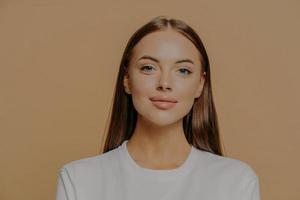 El retrato de una mujer bastante europea con el pelo largo se ve con confianza y la cámara tiene un maquillaje de piel brillante y saludable que lleva un jersey casual aislado sobre una pared marrón del estudio. concepto de belleza y bienestar. foto