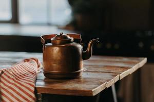 Horizontal shot of aluminium teapot on wooden table against blurred background striped napkin near. Old kettle made of bronze photo