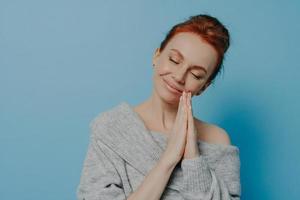 Happy calm redhead grateful woman holding hands in prayer gesture with closed eyes in studio photo