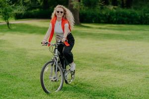 Positive smiling woman dressed in casual clothes, poses on bicycle, has active rest in park, rides on green lawn during sunny summer day, being in high spirit. People, recreation, hobby concept photo