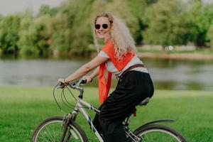 Photo of attractive young woman with curly hair, rides bicycle, wears sunglasses, t shirt, pants, jumper on shoulders, enjoys summer time, fresh air outdoor, spends free time actively with friends