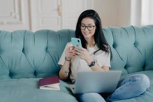 Happy female blogger chats with friends in social netwoks, works on laptop computer, poses in coworking space, sits on sofa in lotus pose, connected to wireless internet. Technology, freelancing photo