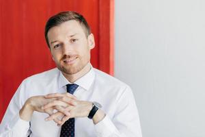 Photo of handsome intelligent man with gentle smile, keeps hands together, wears formal white shirt, poses against red and white background. Male business owner with confident expression. Career