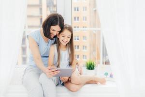 el retrato de una linda mujer abraza a su pequeña hija, ve dibujos animados en una tableta digital, se conecta a internet inalámbrico en casa, se sienta en el alféizar de la ventana con cortinas blancas en casa. niños y padres foto