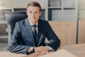 Professional male banker sits in cabinet, wears formal clothes, has confident expression while gives interview concerning banking, poses at office. Man employee arranges meeting of colleagues photo