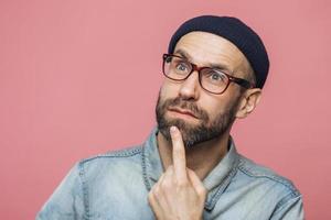 Close up shot of thoughtful bearded male with blue eyes being deep in thoughts, looks pensively into distance, ponders about something, isolated over pink background. Attractive man poses indoor photo