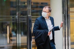 Respiratory protection, coronavirus, dangerous disease. Serious man stands in doors of office building, wears medical mask, holds modern cellphone, newspaper to read article about covid-19 treatment photo
