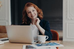 mujer de negocios independiente con cabello de jengibre se sienta frente a la computadora portátil, se comunica con colegas a través de videoconferencia, se sienta en el escritorio, bebe café, tiene una expresión feliz. trabajo a distancia foto