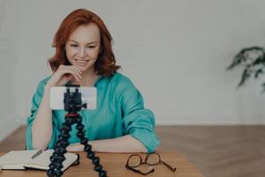 Happy young ginger woman shoots video in front of smartphone videocamera on tripod, gives recommendations how to start own business, has personal vlog, speaks on video call about online training photo