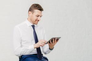 Positive successful male executive manager holds modern touch pad, gets notification about net feeds in social networks, satisfied with high speed internet connection, dressed in formal shirt with tie photo