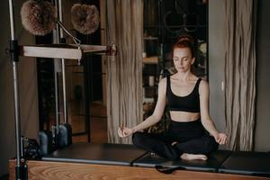 Young redhead woman in sportswear sitting in lotus position on trapeze table trying to relax photo