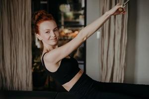 mujer joven de jengibre sonriendo mientras hace ejercicios en reformador de pilates foto