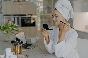 Beautiful young woman applies makeup cosmetics with brush in front of mirror poses against home interior sits at desk with opened laptop and cosmetology tools dressed in bathrobe. Self care concept