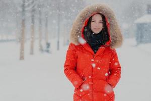 Outdoor shot of attractive woman dressed in winter clothes, keeps both hands in pockets, looks with satisfied expression directly at camera, walks in open air during snowfall. Snowy weather. photo