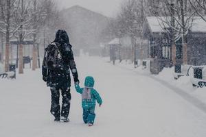 Back view of affectionate father and little male child hold hands, walk along snowy street during heavy snowfall in winter, enjoy recreation time, go home, dressed in warm clothes, carry rucksack photo