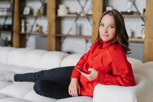 Photo of attractive female model in knitted red sweater, messages online at home, poses on couch in living room, uses modern cell phone and wireless internet connection, looks with appealing look