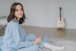 Adorable woman listens lecture on news in internet via cell phone and earphones, has recreation time, connected to wifi at home, poses in bedroom, blurred background with guitar in background photo