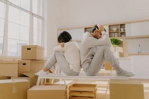 Photo of stressful couple get divorse, leave house, start living seperately, sit backs to each other, pose in kitchen, cardboard boxes with belongings around, focused down with sad expressions