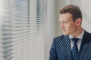 Indoor shot of pensive male economist dressed in formal outfit, looks pensively at window with jalousie conemplates about work during dinner break feels tired after working. Business, people, thoughts photo