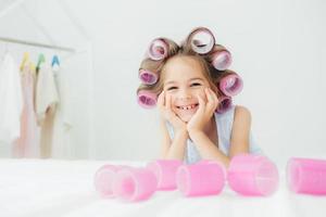 Portrait of cheerful female kid keeps hands under chin, has curlers on hair, going to have nice hairstyle, poses against white background, has charming smile, being in good mood. Children and beauty photo