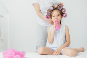 Cropped shot of pleased little child with curlers on head and her mother stands behind combs hair and winds curlers, have appealing appearnce. People, loving family and relationship concept. photo