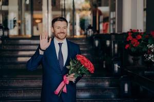 hombre atractivo esperando en la calle con un elegante traje azul oscuro con ramo de rosas rojas foto