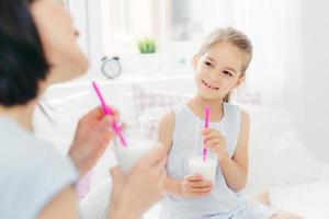 Cropped shot of beautiful small female child drinks tasty milk shake together with her mother, enjoys good morning and calm domestic atmosphere, has happy look. People, breakfast and bedding photo