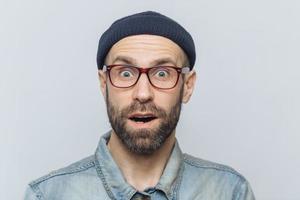 Horizontal shot of amazed excited shocked man stares at camera with unexpected expression, keeps mouth opened, hears awful news, isolated over grey background. Omg and great surprisment concept photo