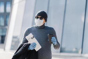 Image of young man wears sterile mask and rubber medical gloves, looks aside, walks through city during infectious disease spread, drinks coffee, folded newspaper. Coronavirus prevention concept photo