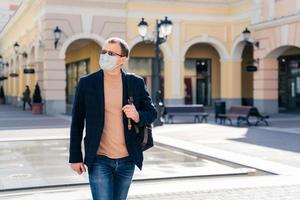 Outdoor shot of serious man walks on station with backpack, concentrated aside, wears medical mask to prevent virus, waits for bus or train, cares about health. Coronavirus pandemic concept. photo