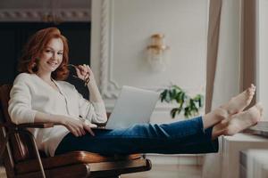 Cheerful successful ginger woman copywriter works from home, keeps laptop on knees, stretches legs, thinks on idea for creating advertising content on own website, browses information in internet photo
