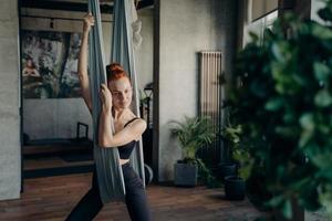 Relaxed red haired woman sitting on aerial yoga hammock and resting after class in fitness studio photo