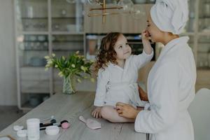 Pretty curly small girl touches nose of her mother dressed in white soft dressing gown sits on table with cosmetic products going to do makeup for mom pose together against cozy home interior photo