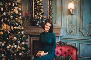 People, celebration, holidays concept. Smiling beautiful woman wears nice dress, sits near Christmas tree in room, holds present, glad to celebrate her favourite holiday. Merry Christmas and New Year